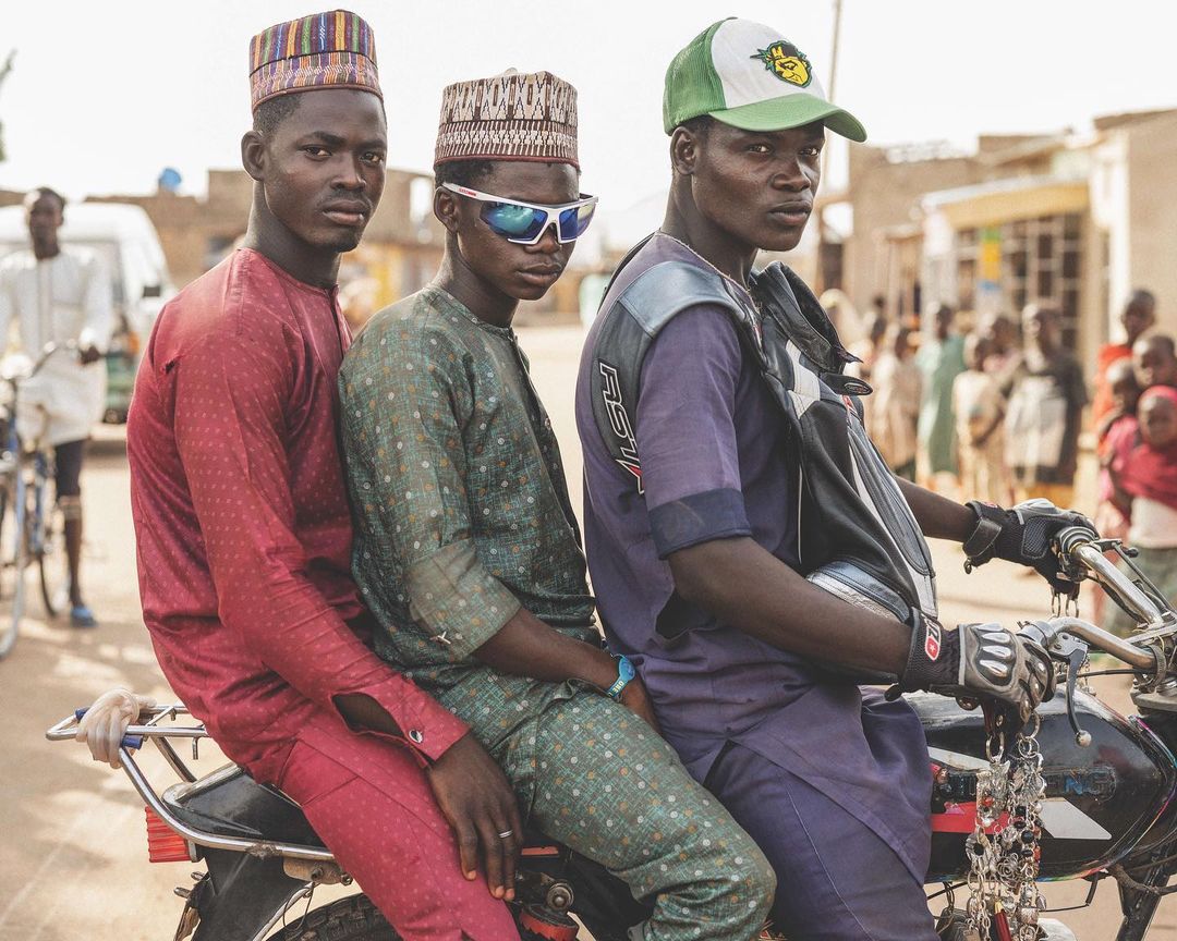 Three young men in traditional caps ride a motorcycle through a rural village, captured in a medium shot with natural daylight.