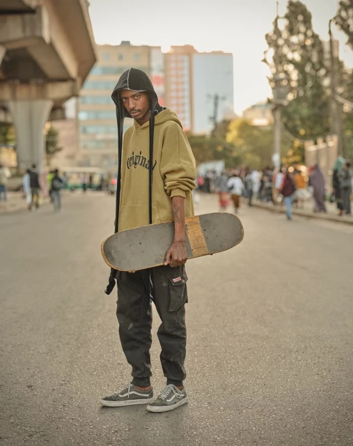 Full-length portrait of a young adult Black man holding a skateboard on a city street with buildings and people in the background.