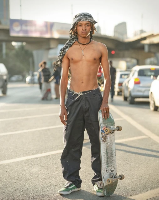 Young man stands shirtless on a city street holding a skateboard, wearing pants, sneakers, and a headscarf with traffic in the background.