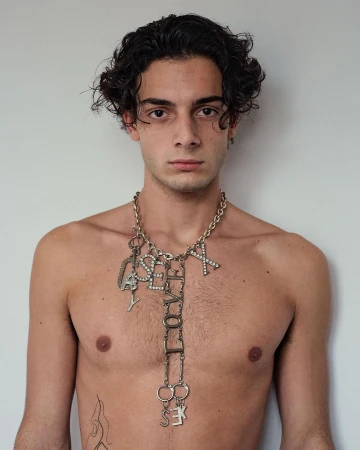 Portrait of a young adult man with dark hair, light skin, wearing a metal 'love, sex, yes' necklace, posing against a plain white background.