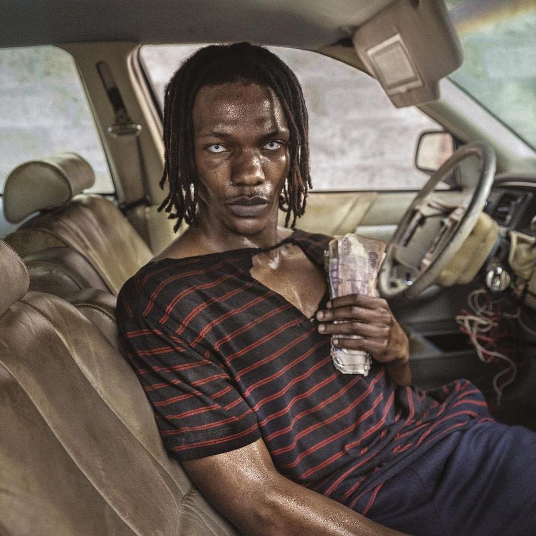Portrait of a Black adult male with pale eyes, holding money inside a car with worn interior, wearing a striped shirt. Close-up.