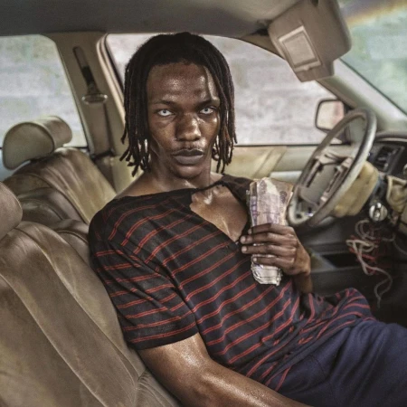 Portrait of a Black adult male with pale eyes, holding money inside a car with worn interior, wearing a striped shirt. Close-up.