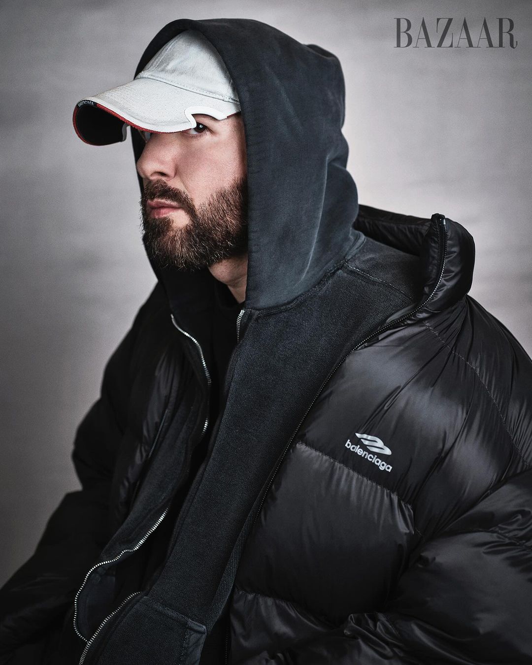 Portrait of adult male with beard wearing black hooded puffer jacket and baseball cap in front of grey studio backdrop, Bazaar logo.
