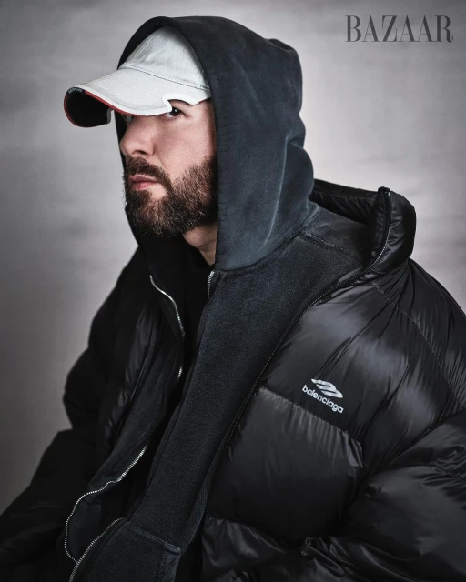 Portrait of adult male with beard wearing black hooded puffer jacket and baseball cap in front of grey studio backdrop, Bazaar logo.