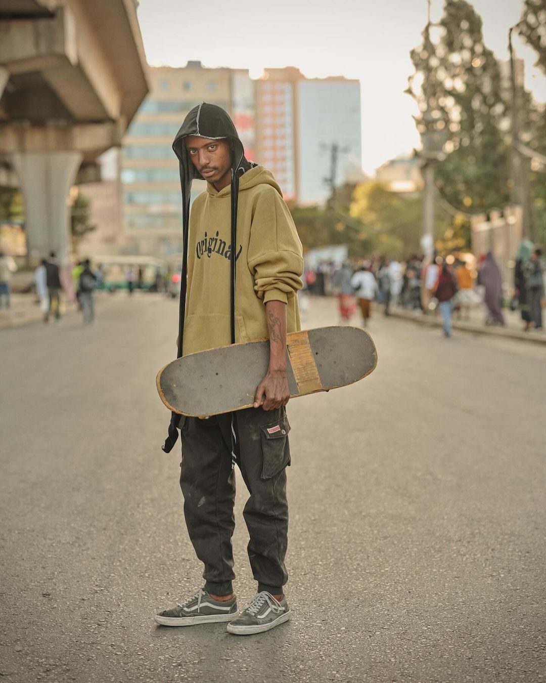 A young adult with a skateboard stands on a city street under an overpass, wearing a hooded sweatshirt, cargo pants, and sneakers on a sunny day.