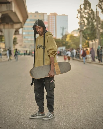 A young adult with a skateboard stands on a city street under an overpass, wearing a hooded sweatshirt, cargo pants, and sneakers on a sunny day.