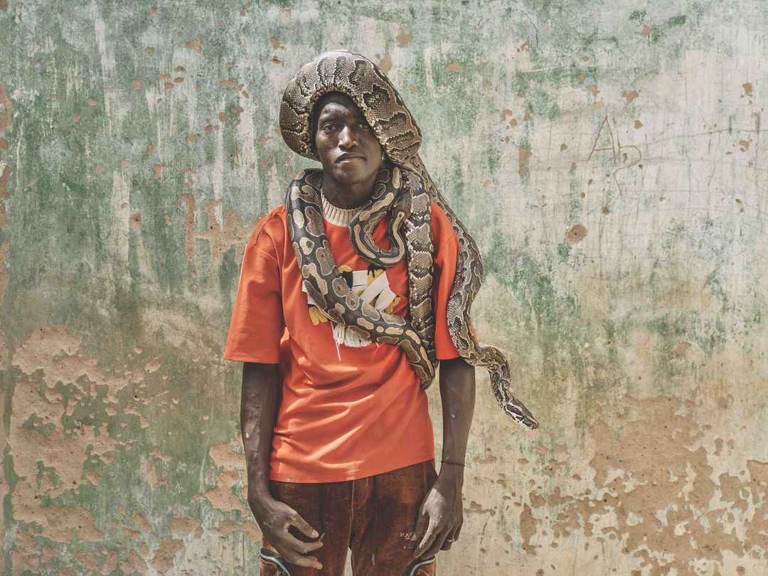 An African man stands outdoors with a large snake wrapped around his head and shoulders in a portrait against a weathered wall.