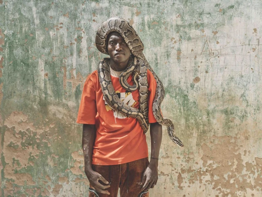 An African man stands outdoors with a large snake wrapped around his head and shoulders in a portrait against a weathered wall.