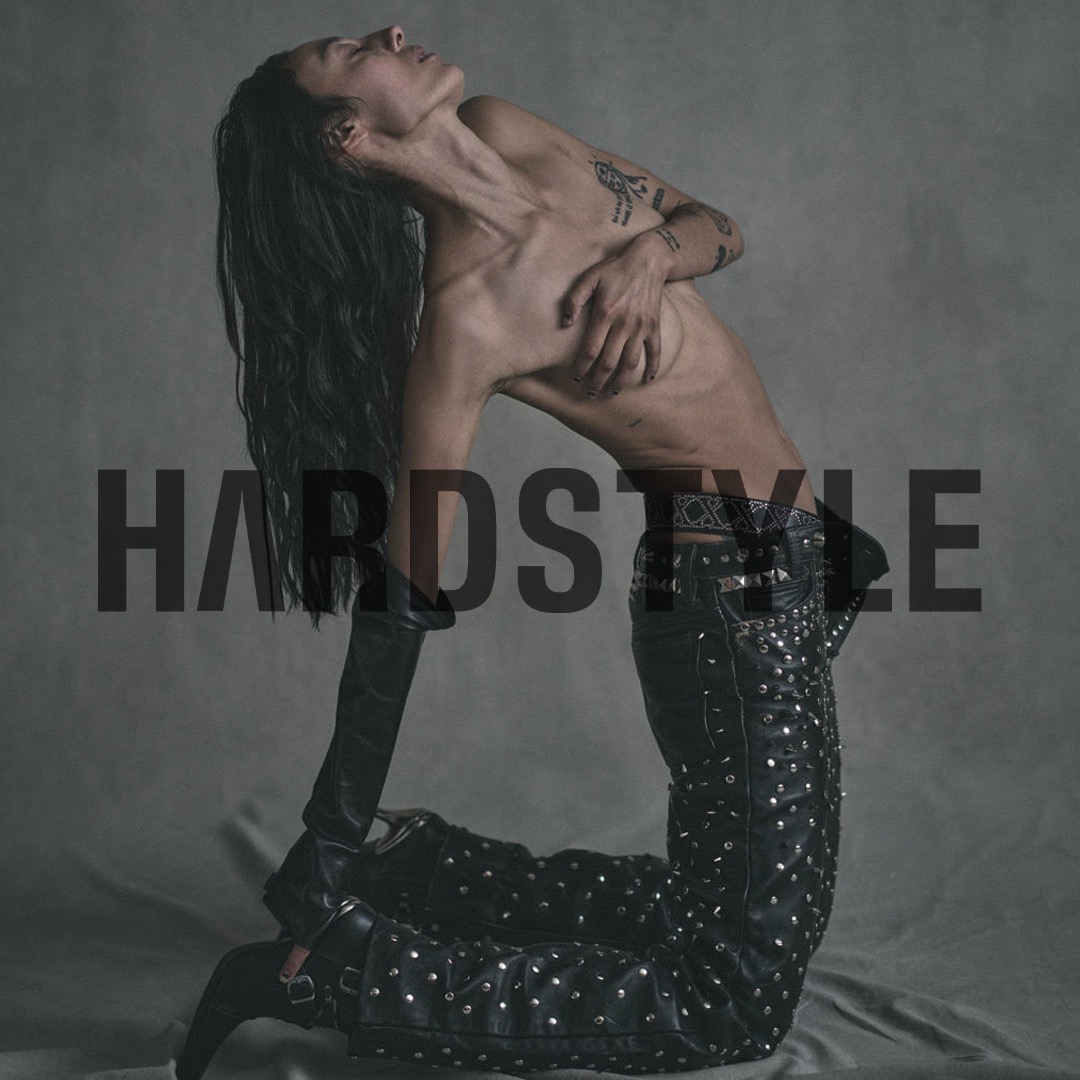 A person with long hair poses kneeling, wearing black studded pants, elbow-length gloves, covering their chest, against a grey backdrop.