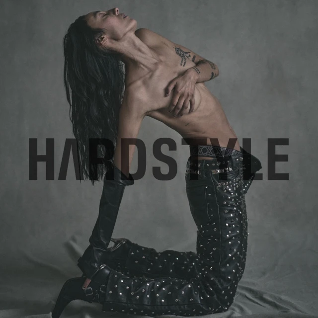A person with long hair poses kneeling, wearing black studded pants, elbow-length gloves, covering their chest, against a grey backdrop.