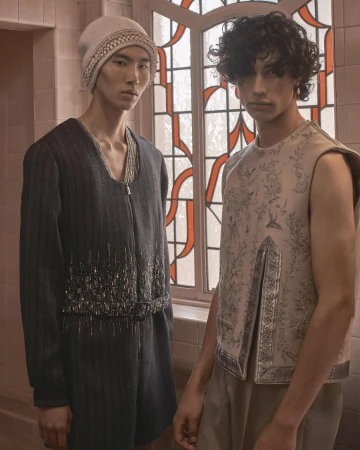Two men pose indoors near a window, showcasing detailed sleeveless and sleeved tops in a fashion editorial setting with neutral tones.