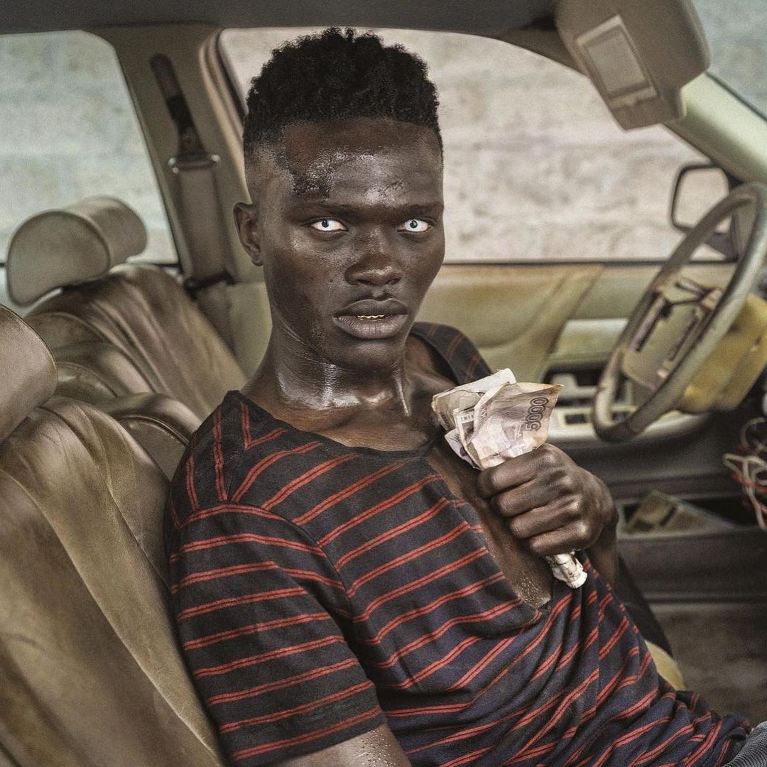 Adult male with dark skin sits in a car seat with stack of money. He is wearing a striped shirt with red and black stripes.