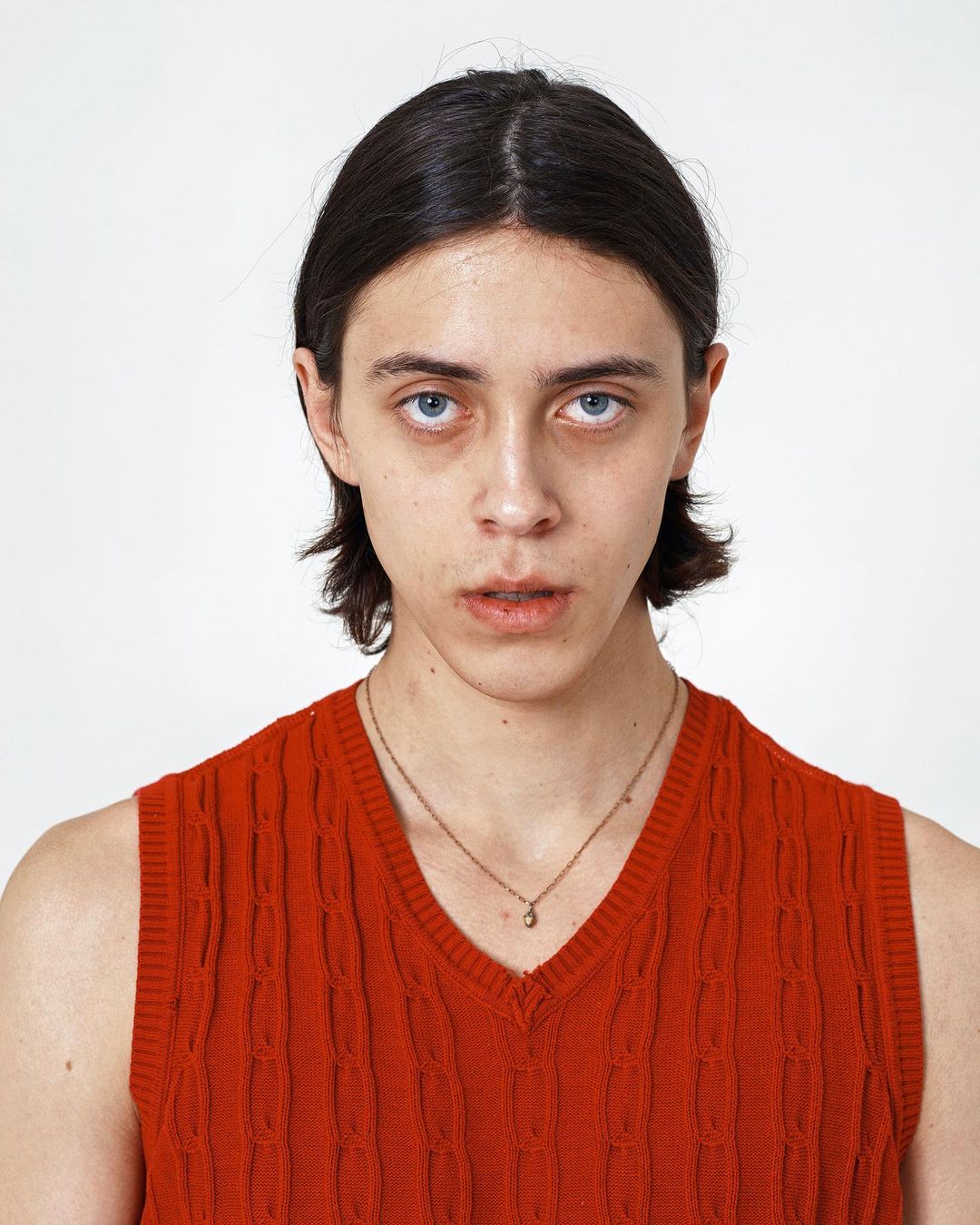 Close-up portrait of a fair-skinned adult with dark hair wearing an orange sweater vest against a plain white backdrop.