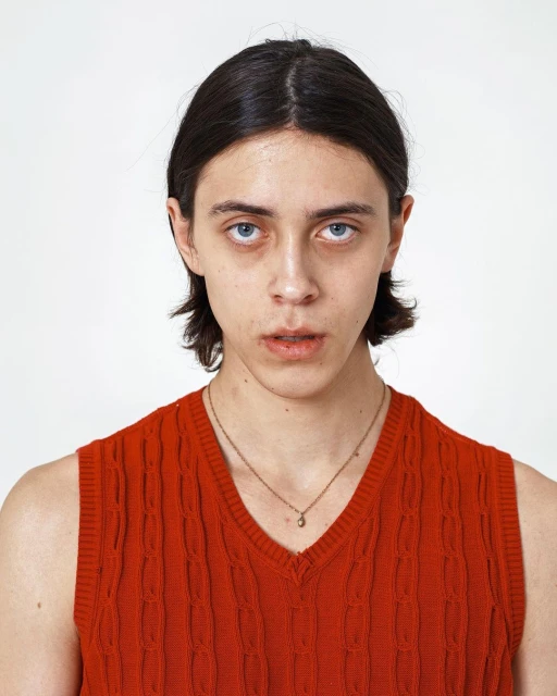 Close-up portrait of a fair-skinned adult with dark hair wearing an orange sweater vest against a plain white backdrop.