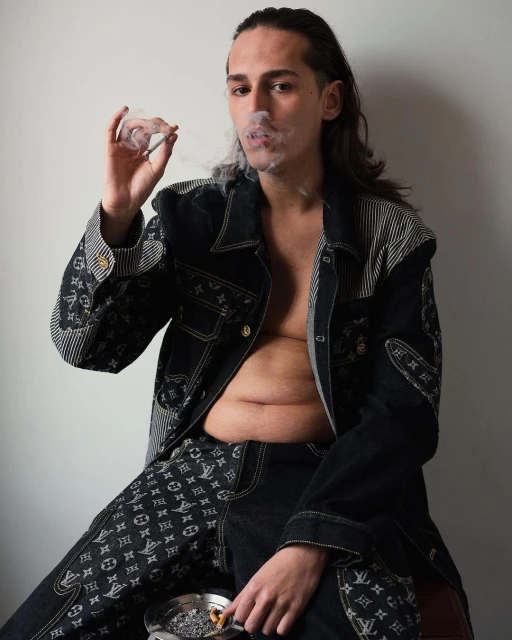 Person smoking indoors wearing a patterned denim jacket and pants. An ashtray is also visible. Dark hair and neutral background.