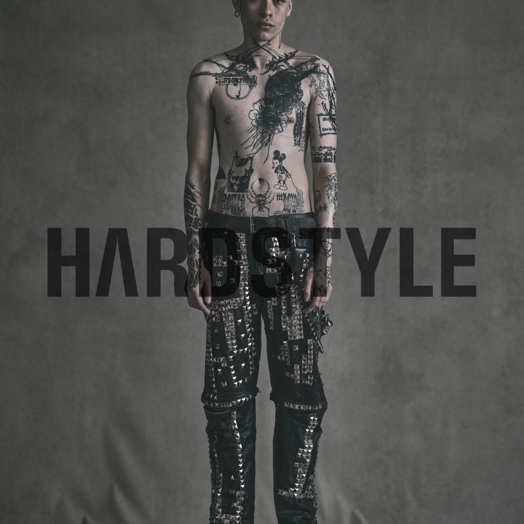 Man with tattoos stands in studded pants against a neutral backdrop in studio lighting. Fashion and portrait photography.