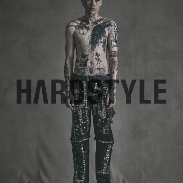 Man with tattoos stands in studded pants against a neutral backdrop in studio lighting. Fashion and portrait photography.
