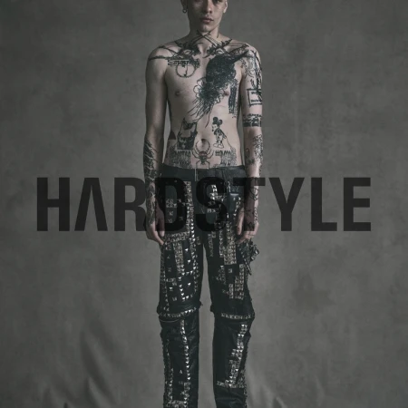 Man with tattoos stands in studded pants against a neutral backdrop in studio lighting. Fashion and portrait photography.