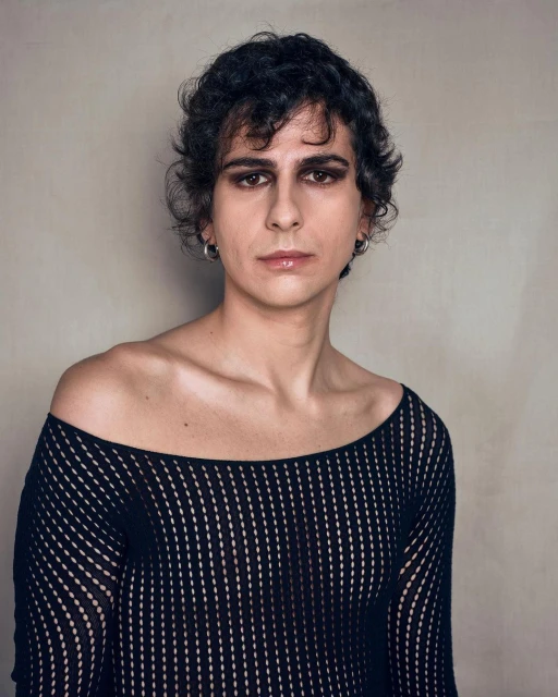Portrait of a person with curly dark hair wearing a black textured top against a neutral backdrop. Eye-level shot, indoors.