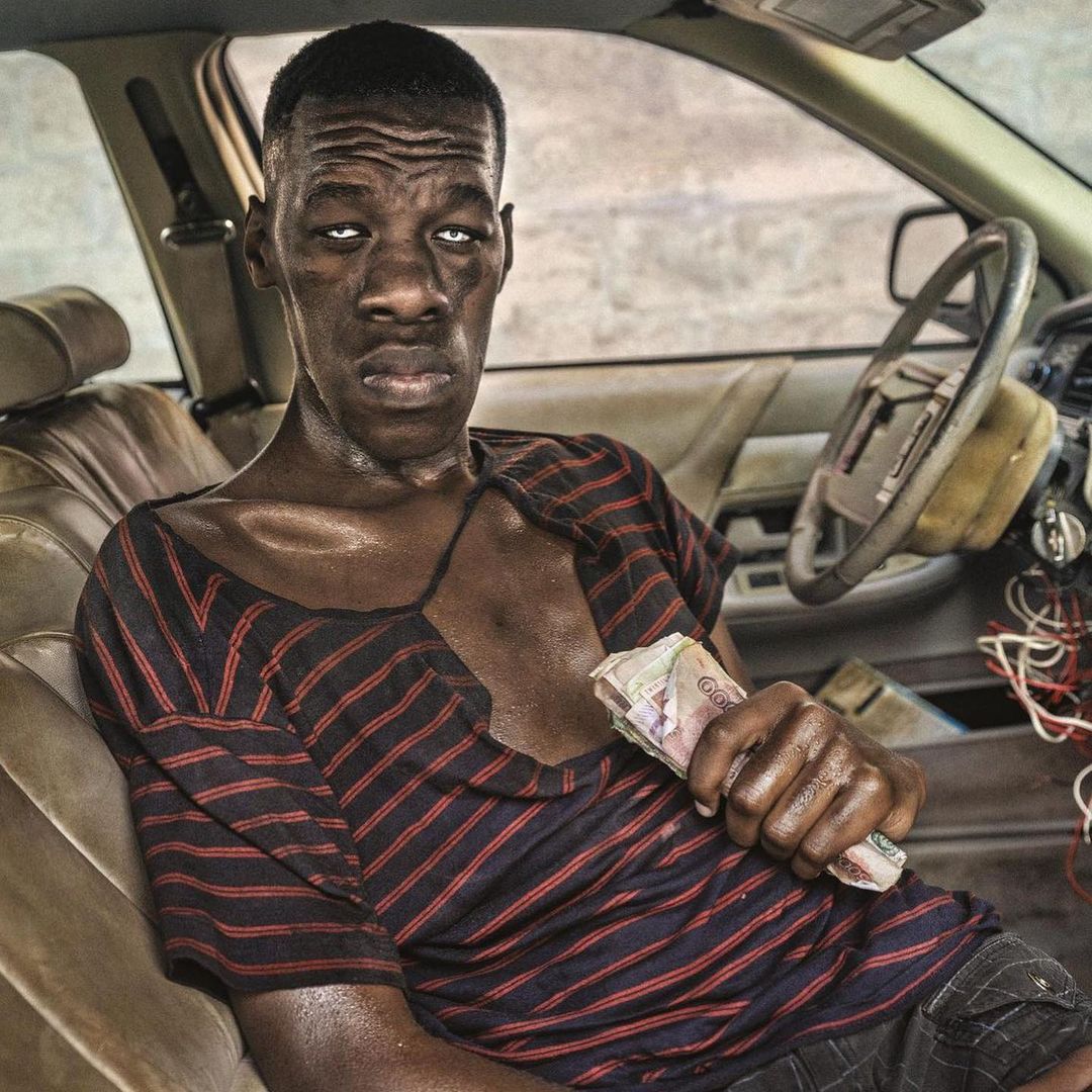 Adult male seated in a worn car interior, holding a stack of cash, wearing a striped shirt, with a serious expression. 