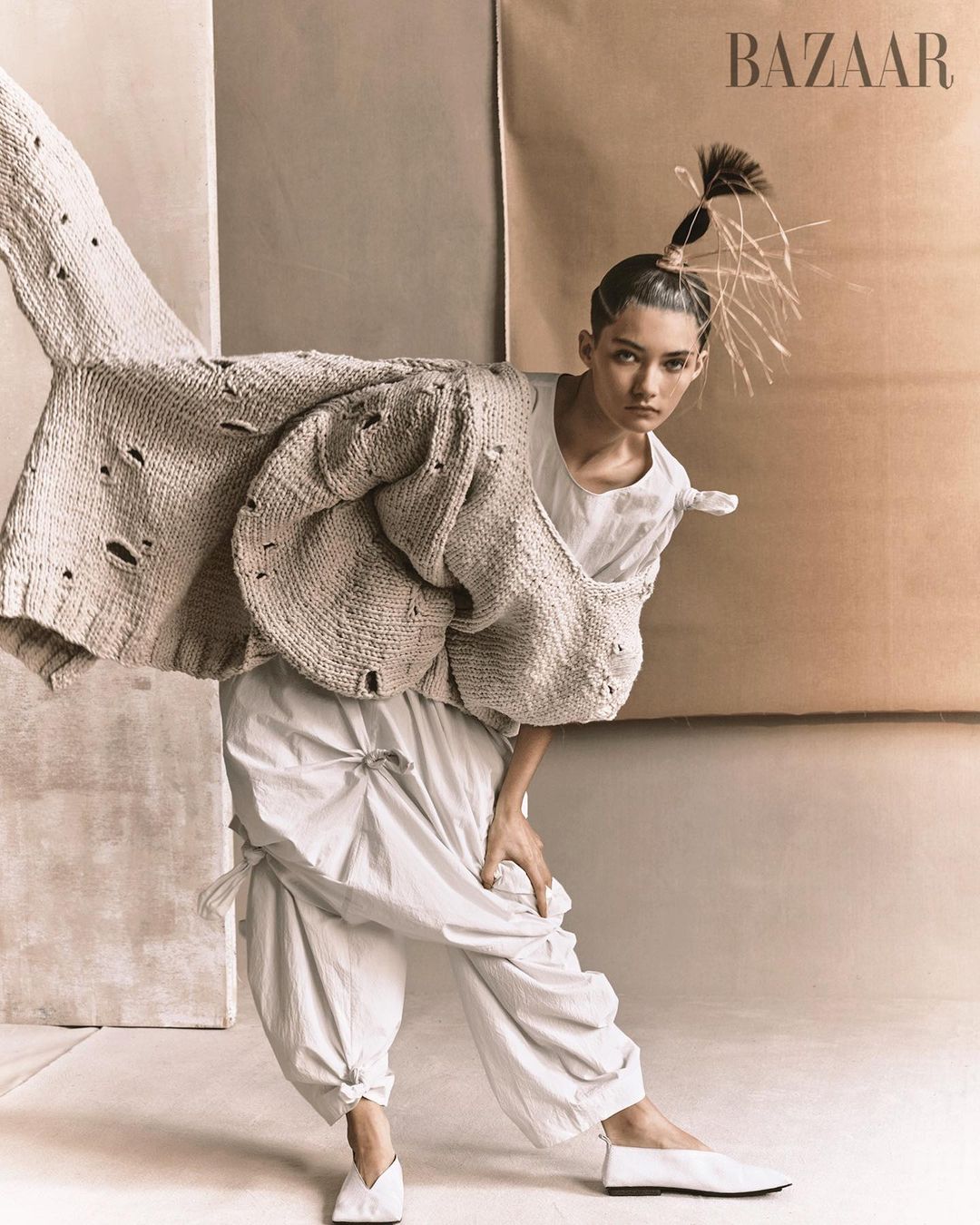 Editorial fashion shot of a woman in a white outfit and knit cardigan posing in front of neutral-toned paper backdrops in a studio.