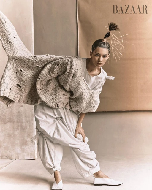 Editorial fashion shot of a woman in a white outfit and knit cardigan posing in front of neutral-toned paper backdrops in a studio.