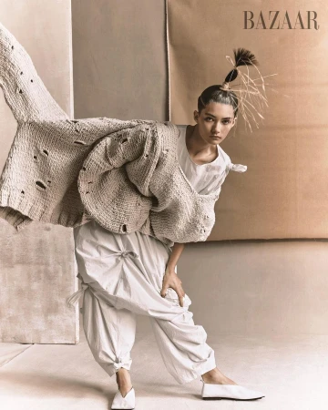 Editorial fashion shot of a woman in a white outfit and knit cardigan posing in front of neutral-toned paper backdrops in a studio.