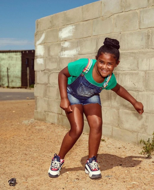 A young African American girl poses outside in a denim romper and sneakers next to a concrete block wall and sandy ground on a sunny day.