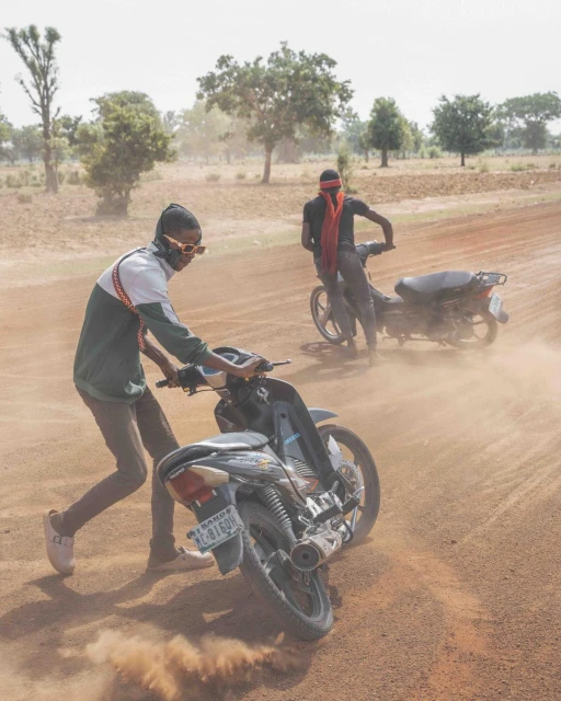 Two men ride motorcycles on a dusty dirt road in a rural area, with trees in the background and a hazy sky overhead during daytime.