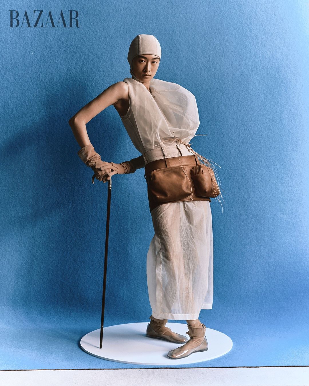 Editorial fashion image of a slender adult woman in sheer white dress with sculptural details holding walking stick against blue backdrop.