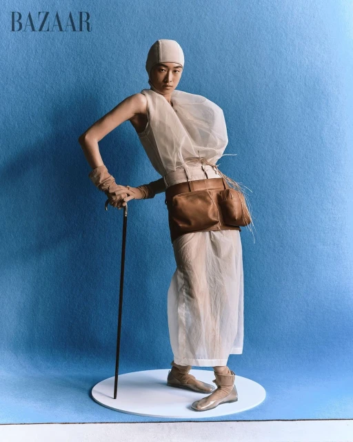 Editorial fashion image of a slender adult woman in sheer white dress with sculptural details holding walking stick against blue backdrop.