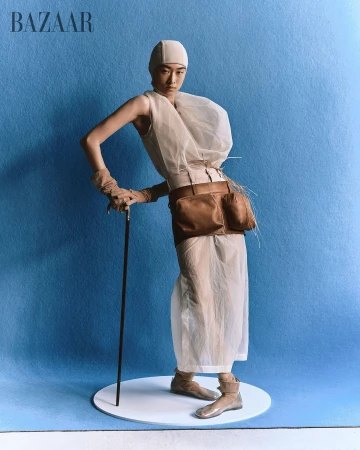 Editorial fashion image of a slender adult woman in sheer white dress with sculptural details holding walking stick against blue backdrop.
