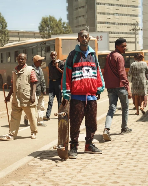 Young man stands with a skateboard on an urban street, surrounded by pedestrians and a bus, on a sunny day in the city.