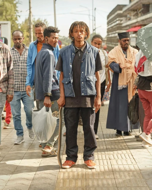 A young adult stands on a city street surrounded by a diverse group of pedestrians, holding a skateboard in an eye-level, full shot.