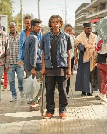 A young adult stands on a city street surrounded by a diverse group of pedestrians, holding a skateboard in an eye-level, full shot.