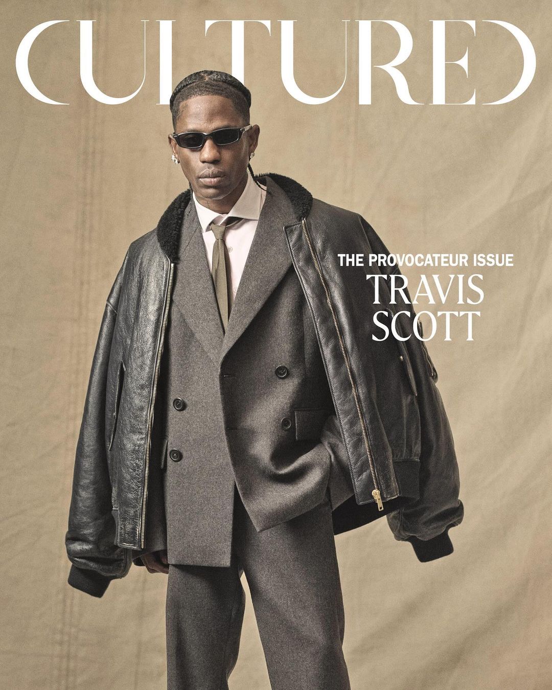 Travis Scott on the cover of Cultured Magazine wearing a gray suit, sunglasses, and black bomber jacket with a neutral background.