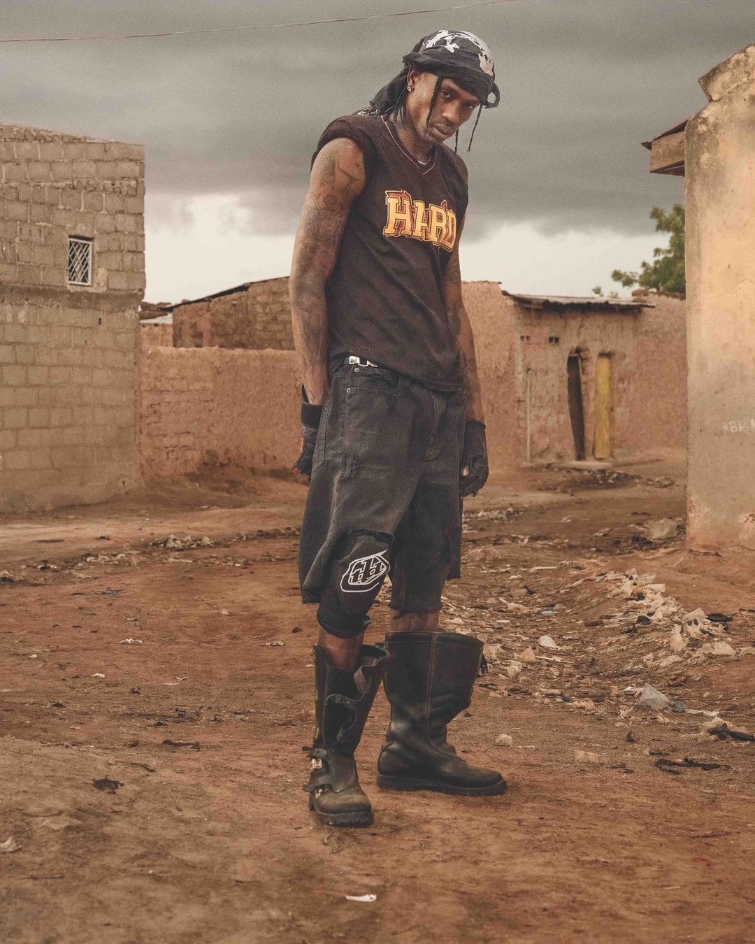 Man with tattoos and head covering poses outdoors in black sleeveless shirt, shorts, and boots, standing in front of simple buildings.