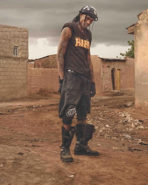 Man with tattoos and head covering poses outdoors in black sleeveless shirt, shorts, and boots, standing in front of simple buildings.