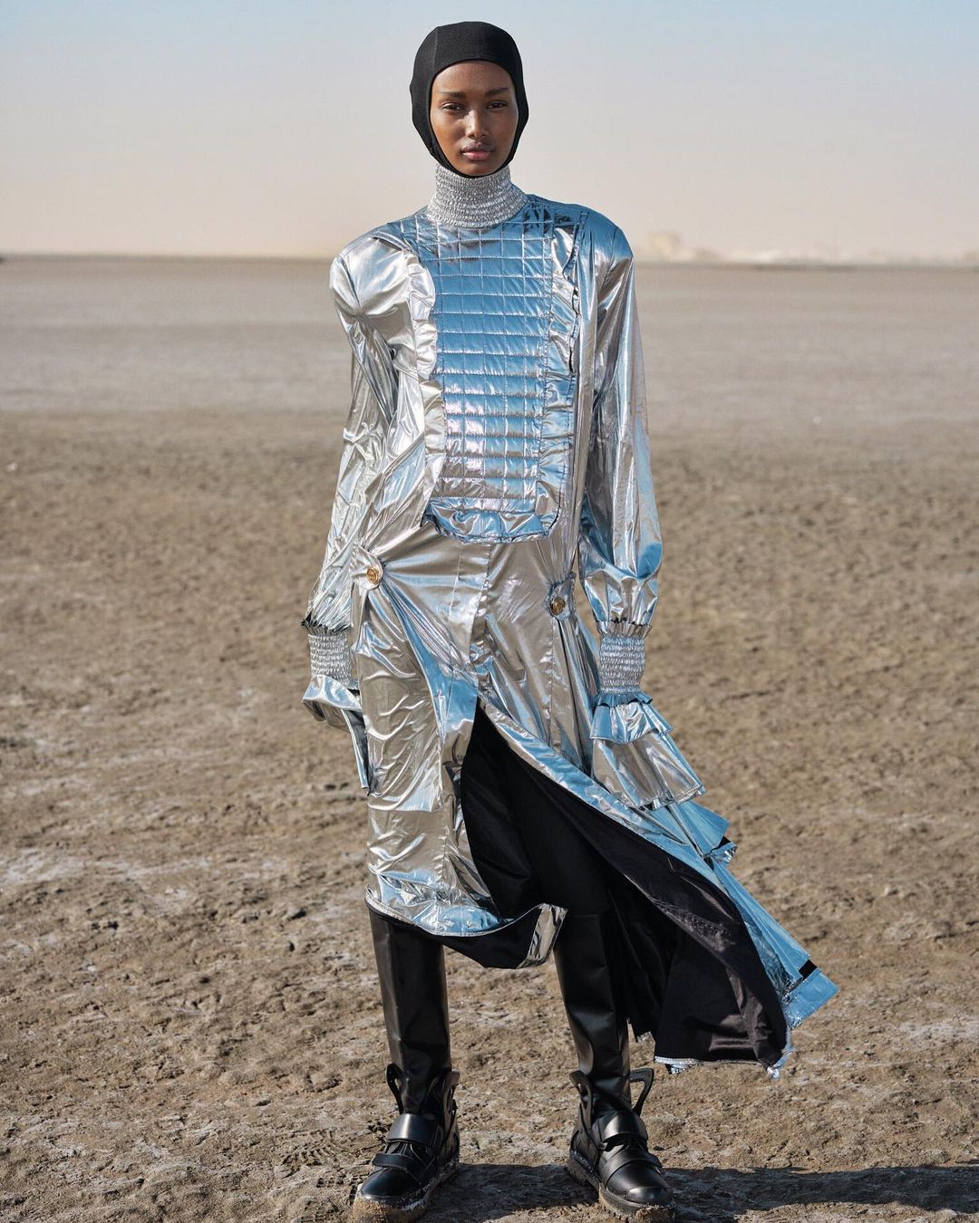 Fashion model in a silver metallic outfit with black boots poses in a barren desert landscape under a clear sky for a fashion shoot.