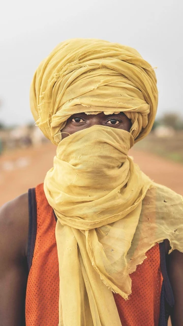 Portrait of an African man with a yellow turban covering his face, standing outdoors on a dirt road, looking directly at the camera.