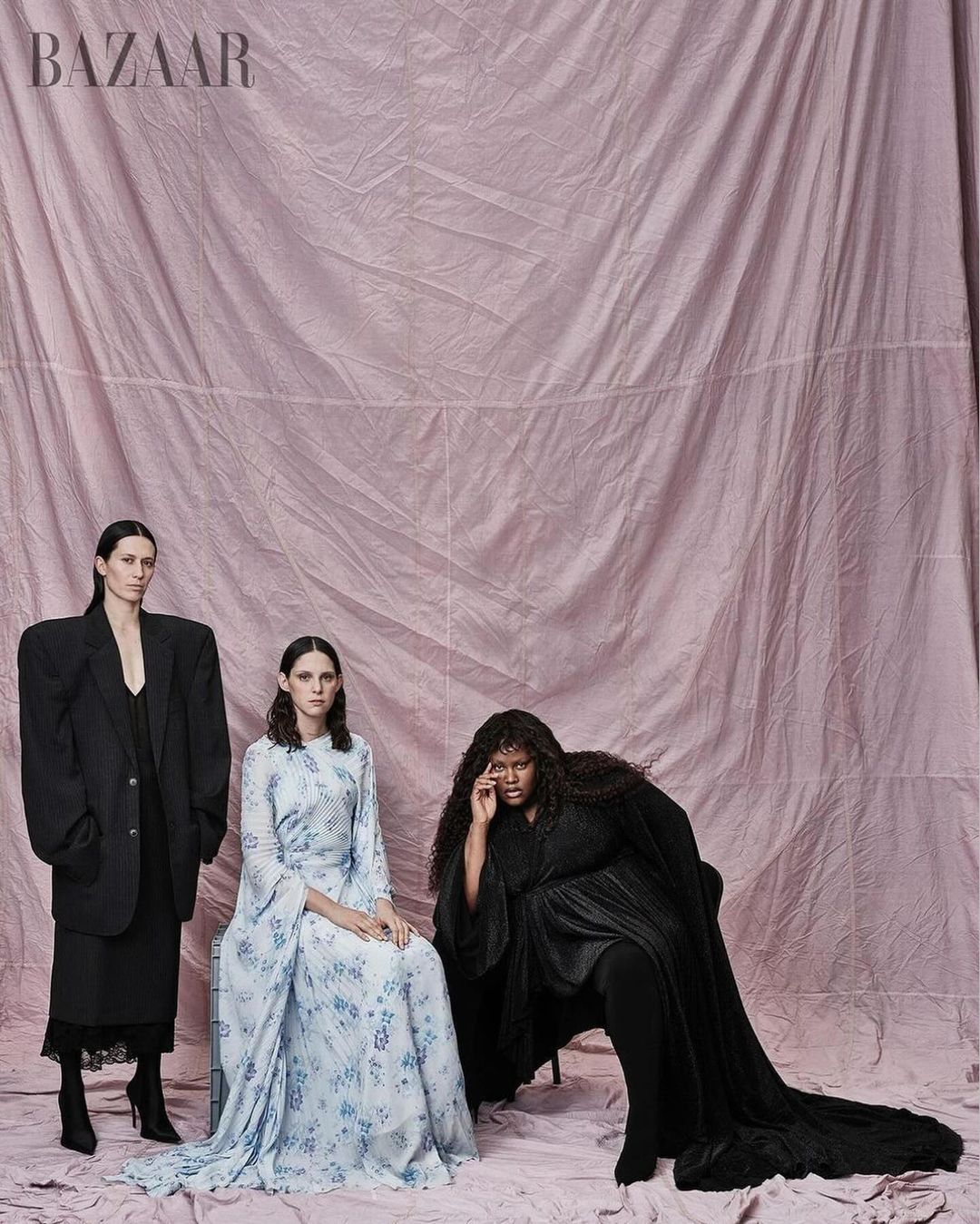 Three fashion models pose in an indoor studio setting against a blush pink backdrop, showcasing unique and elegant dress and suit styles.