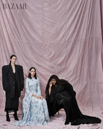 Three fashion models pose in an indoor studio setting against a blush pink backdrop, showcasing unique and elegant dress and suit styles.