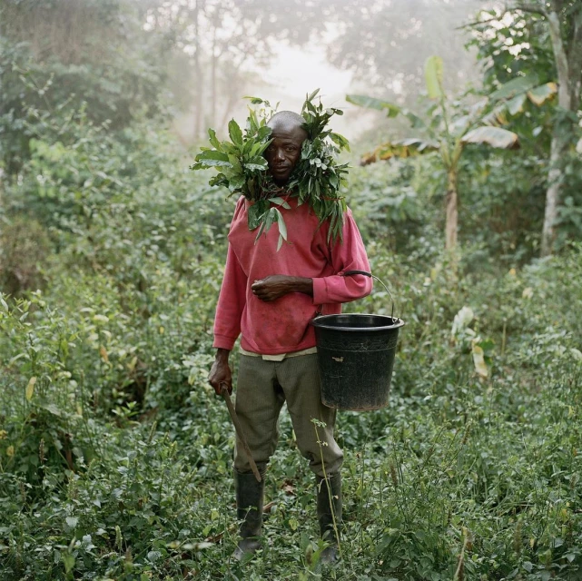 African man stands in lush jungle, adorned with green leaves, carrying a black bucket, amidst vibrant greenery and misty atmosphere.