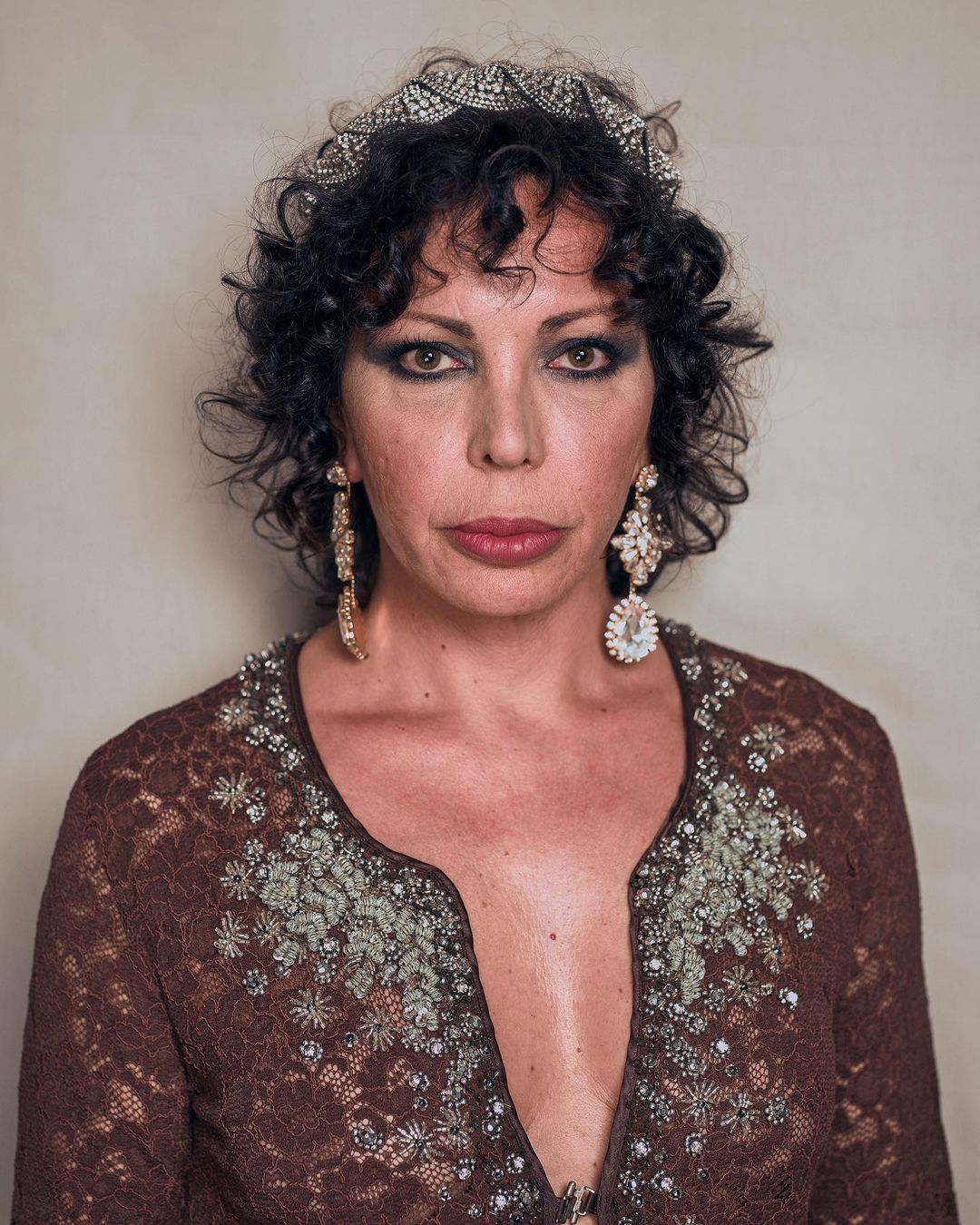 Portrait of an adult woman with curly dark hair wearing a brown floral lace top, large chandelier earrings, and a jeweled tiara.