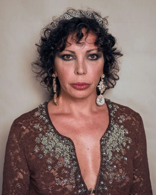 Portrait of an adult woman with curly dark hair wearing a brown floral lace top, large chandelier earrings, and a jeweled tiara.