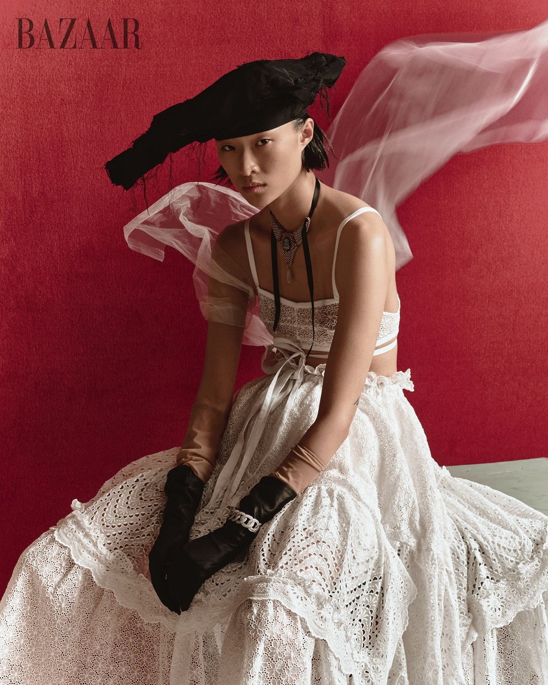 Asian woman in haute couture fashion, wearing a large black hat and lace dress, sitting against a red background in studio lighting.
