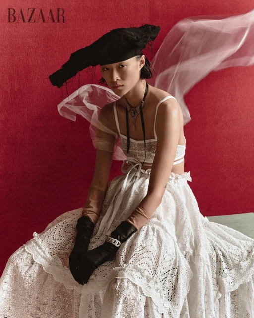 Asian woman in haute couture fashion, wearing a large black hat and lace dress, sitting against a red background in studio lighting.