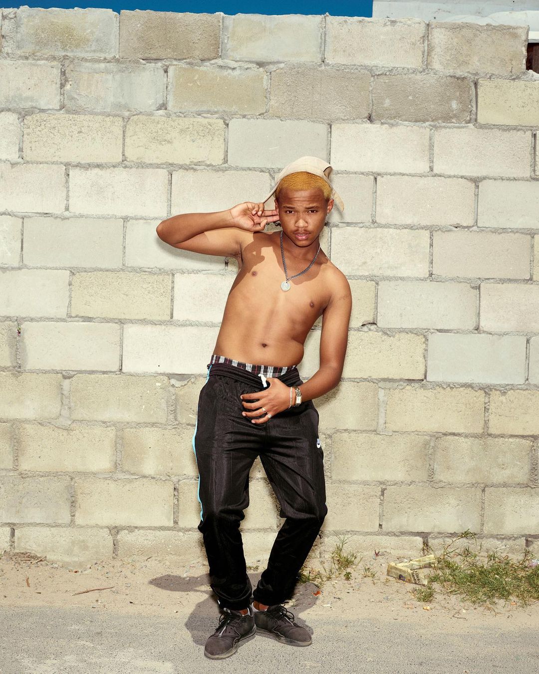 Young adult man with bleached blond hair poses shirtless outdoors by a brick wall with black track pants and dark sneakers.