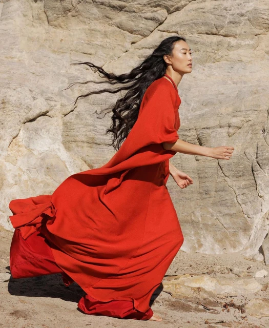 Woman in flowing red dress walking on beach with rock formations in background; long dark hair, fashion photography.