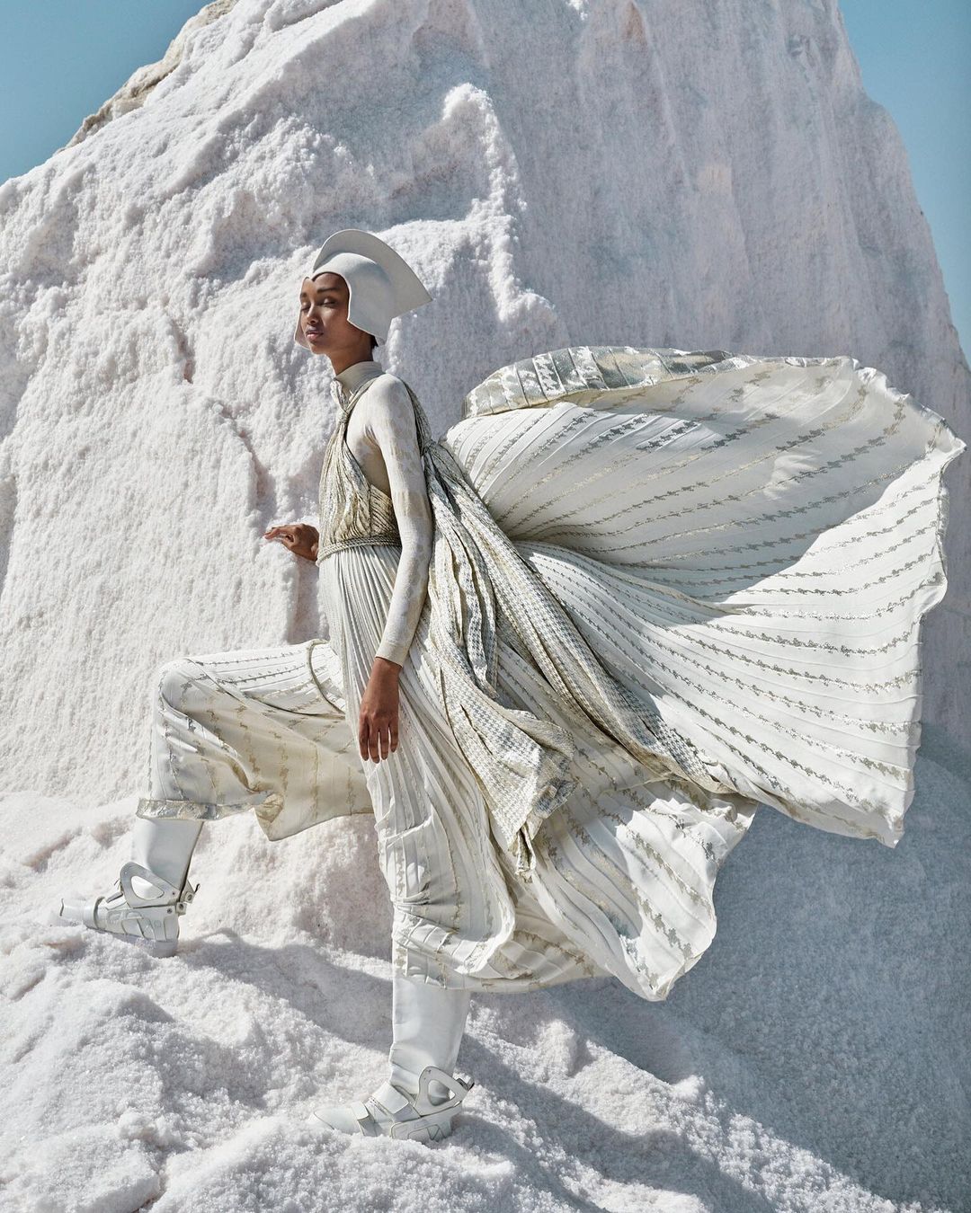 Fashion editorial shot of a woman in a white couture outfit and headpiece posing on a stark white textured salt flat landscape.